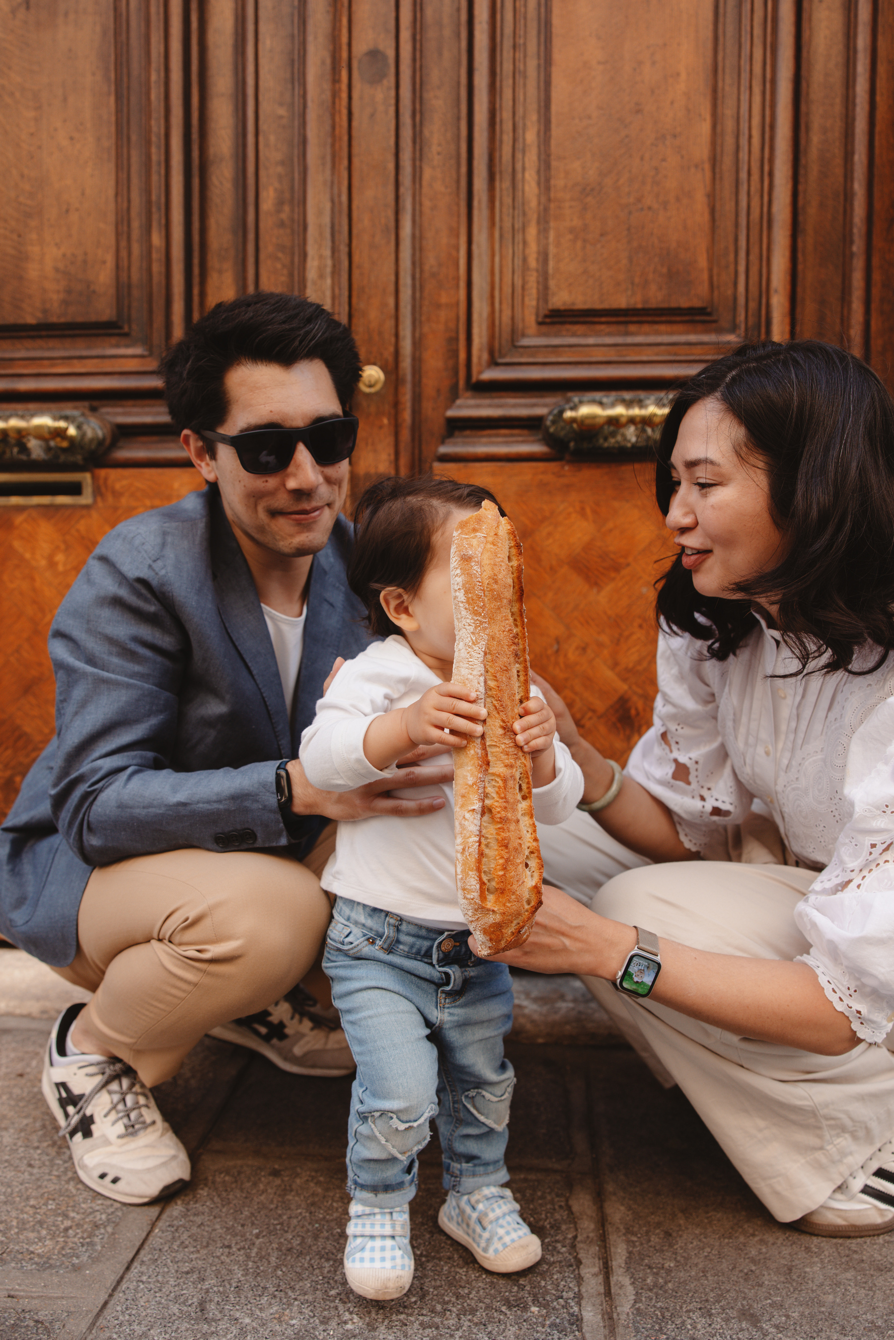 Auto-generated description: A family with a young child holding a large baguette stands in front of wooden doors, with the adults wearing sunglasses and casual clothing.