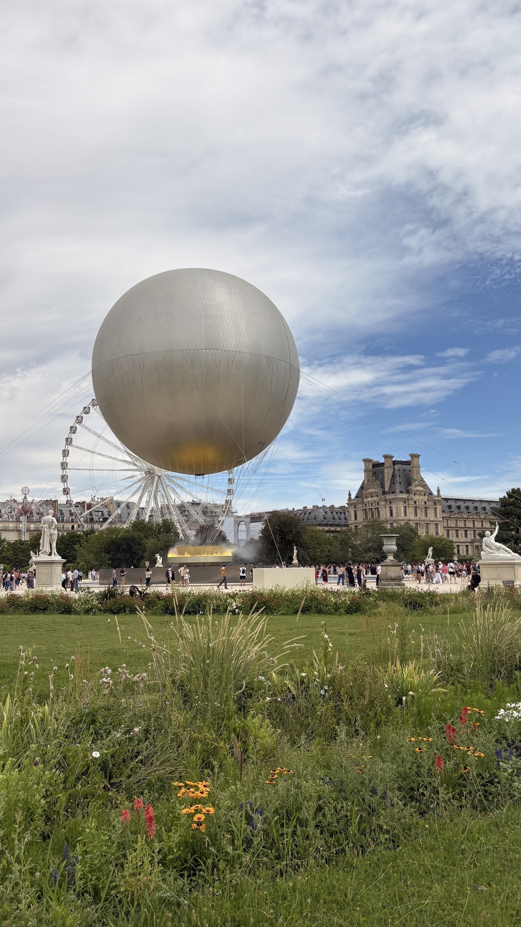 Auto-generated description: A large balloon hovers above a garden with colorful flowers, surrounded by people and historical architecture in the background.