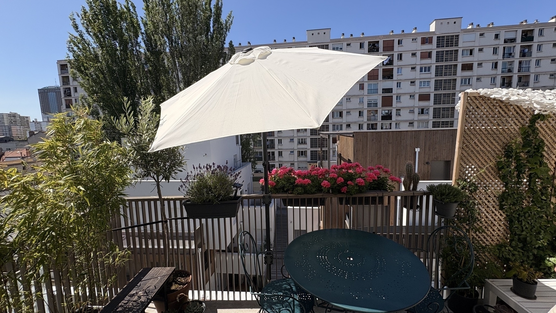 Auto-generated description: A rooftop terrace features vibrant pink flowers, a white umbrella, green plants, and overlooks a cityscape with a large residential building.