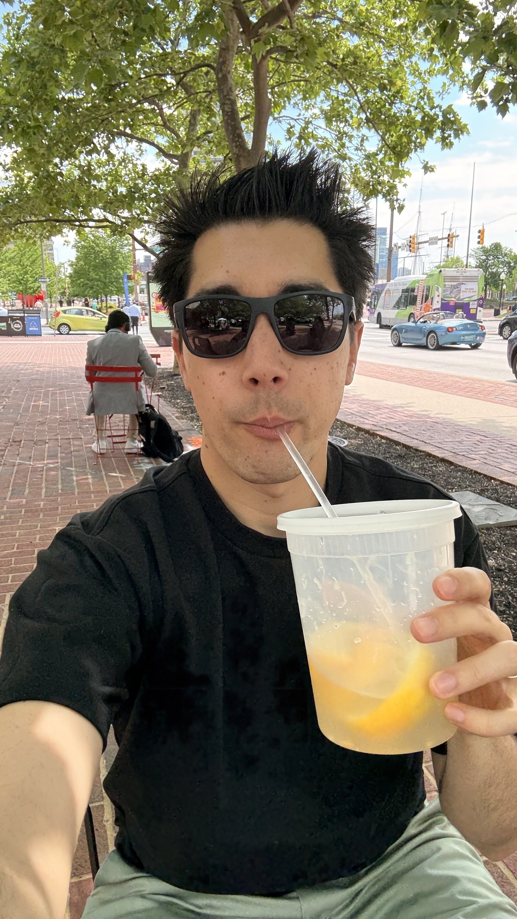 Auto-generated description: A person wearing sunglasses is sitting outdoors, drinking from a large container with a straw.