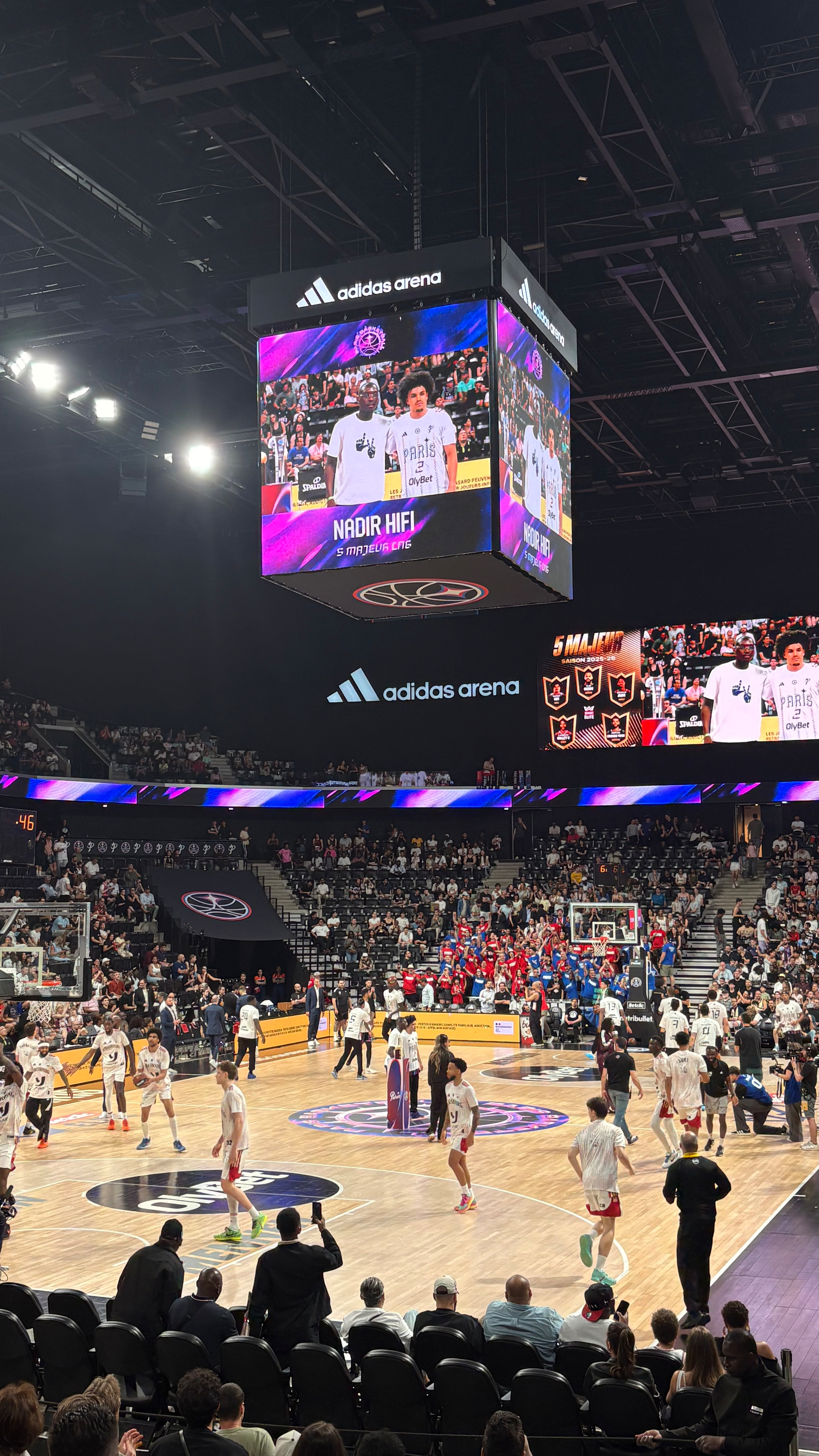 Auto-generated description: A basketball game is taking place in an indoor arena with players warming up on the court and spectators watching from the stands.
