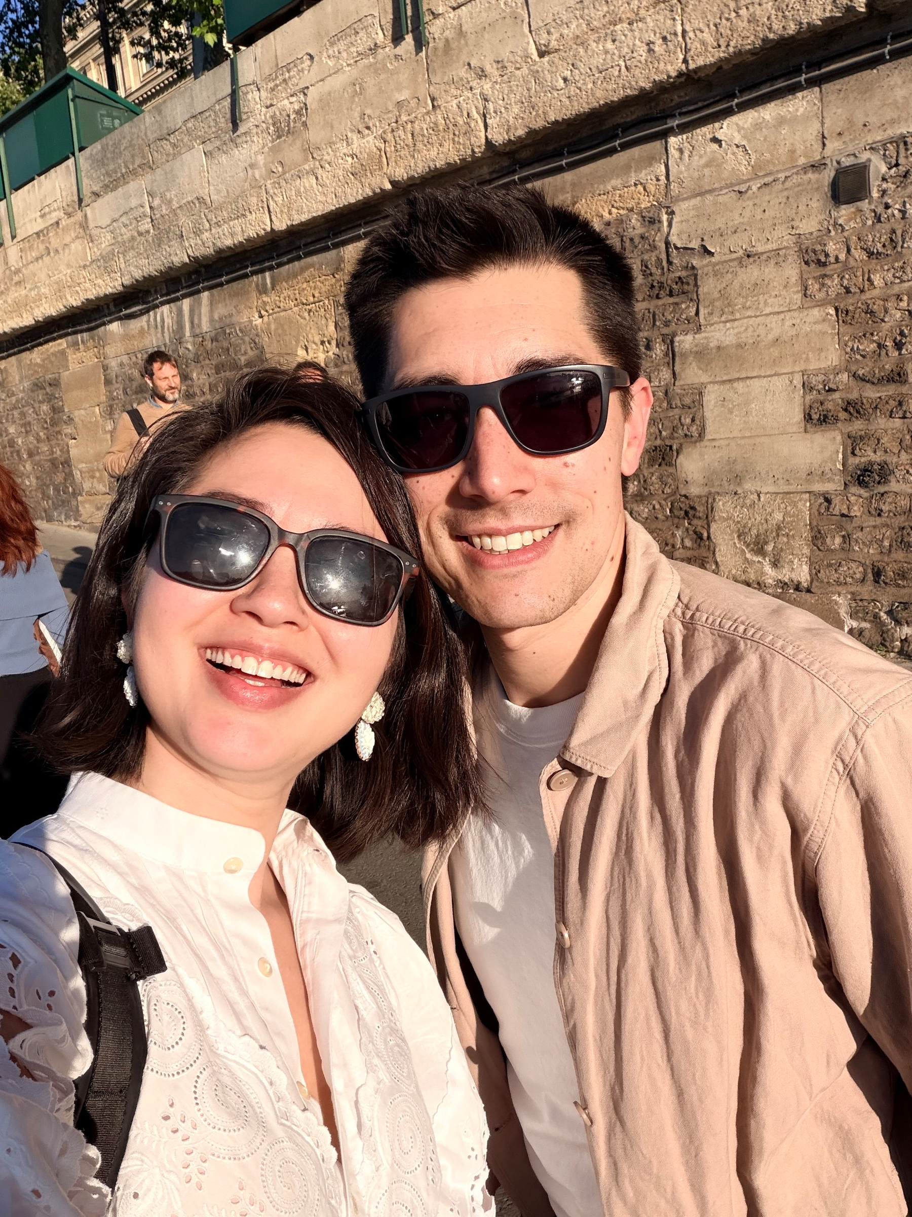Auto-generated description: A smiling couple wearing sunglasses enjoys a sunny day in front of a stone wall.