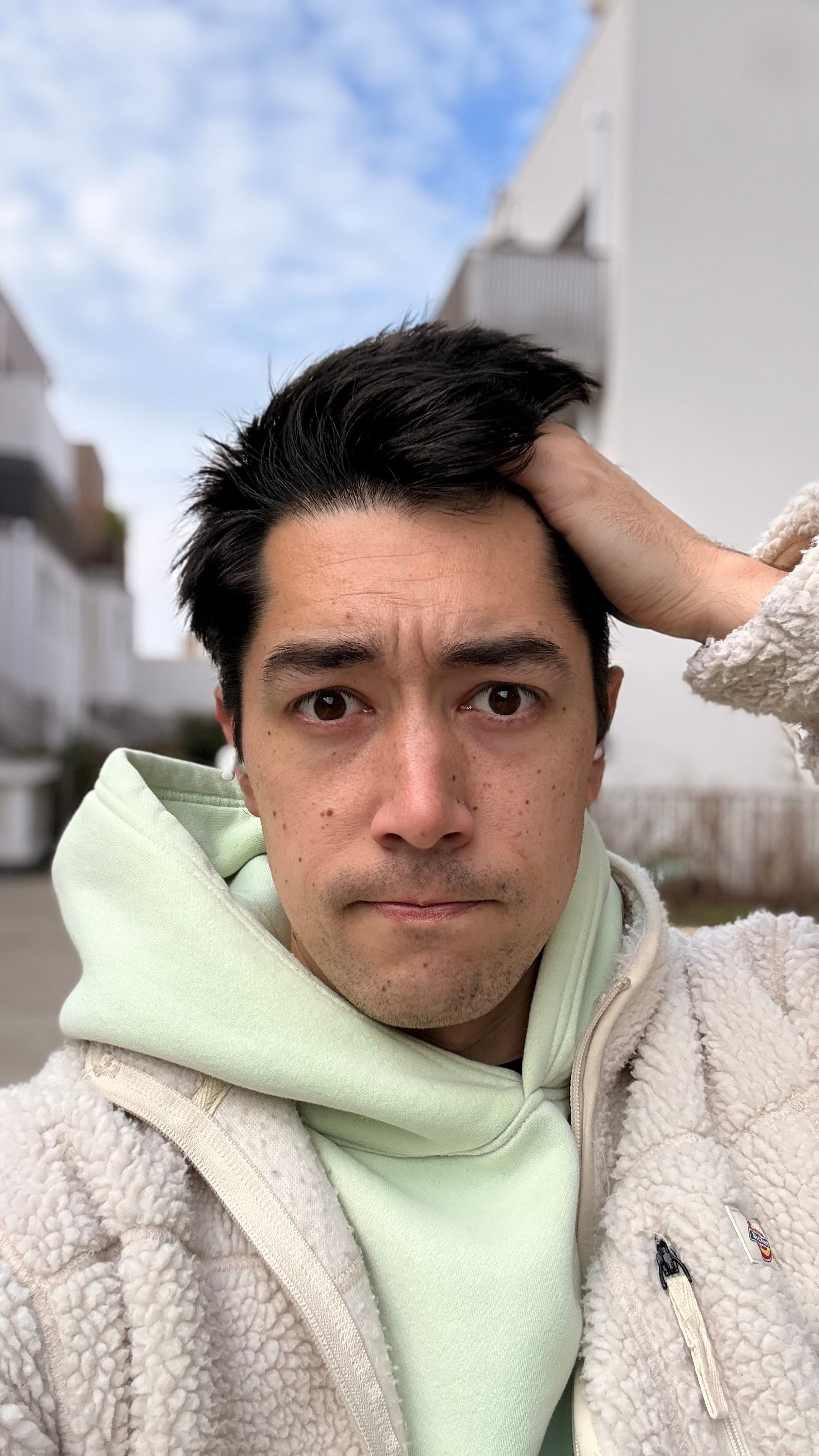 Auto-generated description: A person wearing a light green hoodie and a fluffy jacket stands outdoors, slightly frowning and holding their hair with one hand.