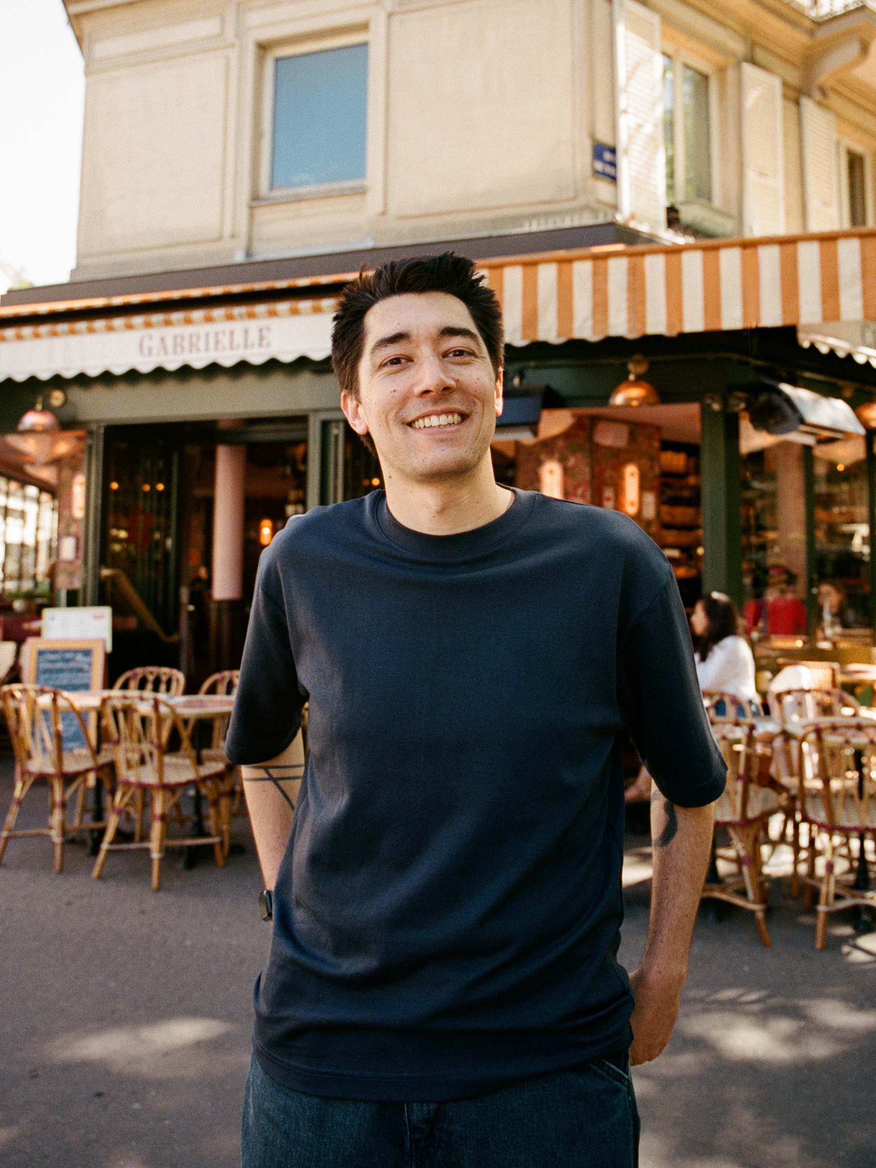 Auto-generated description: A smiling young man with dark hair is standing casually in front of a quaint café with outdoor seating.