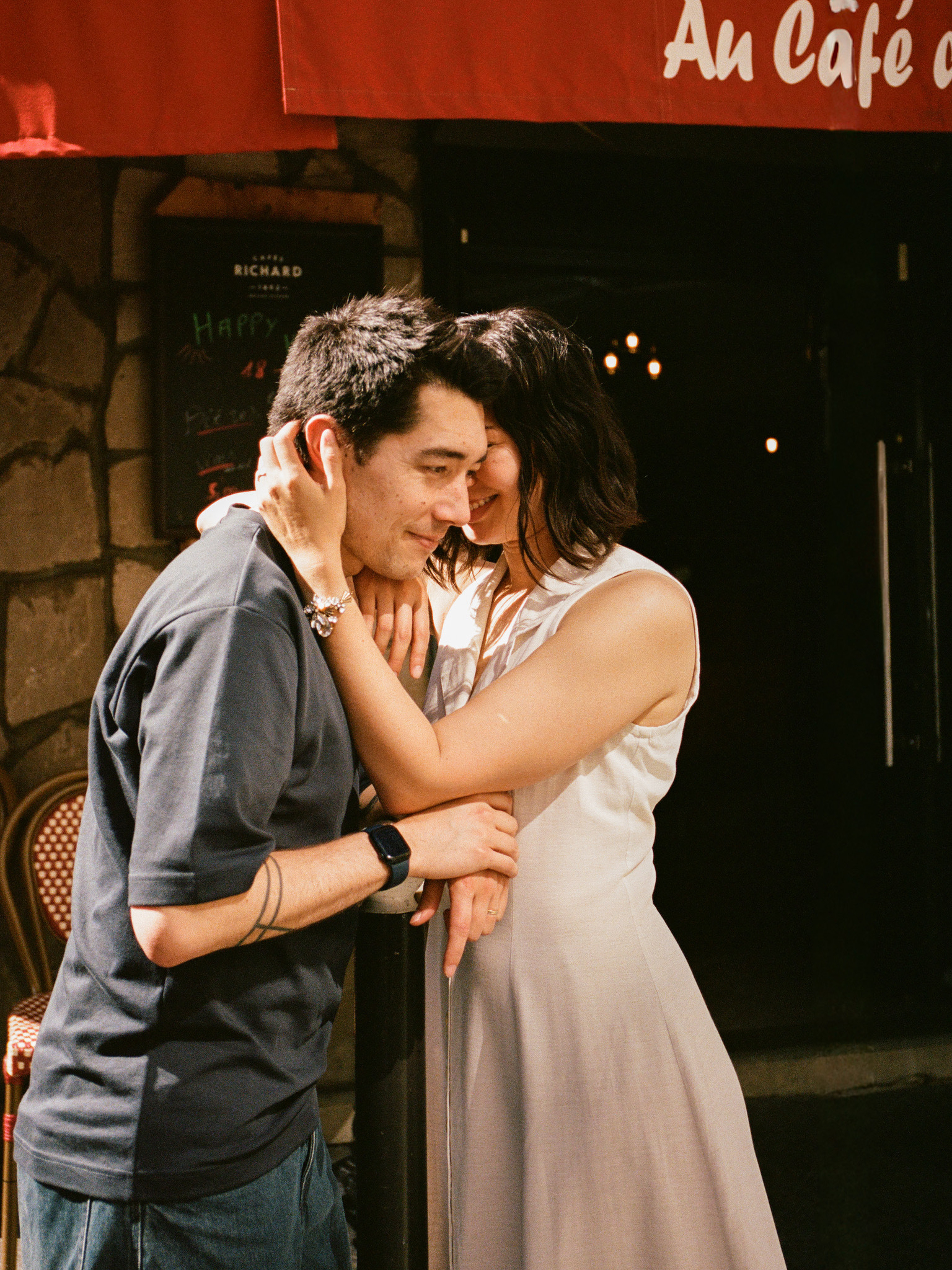 Auto-generated description: A couple shares an intimate and joyful moment outside a café, with the woman gently holding the man's face as they smile warmly at each other.