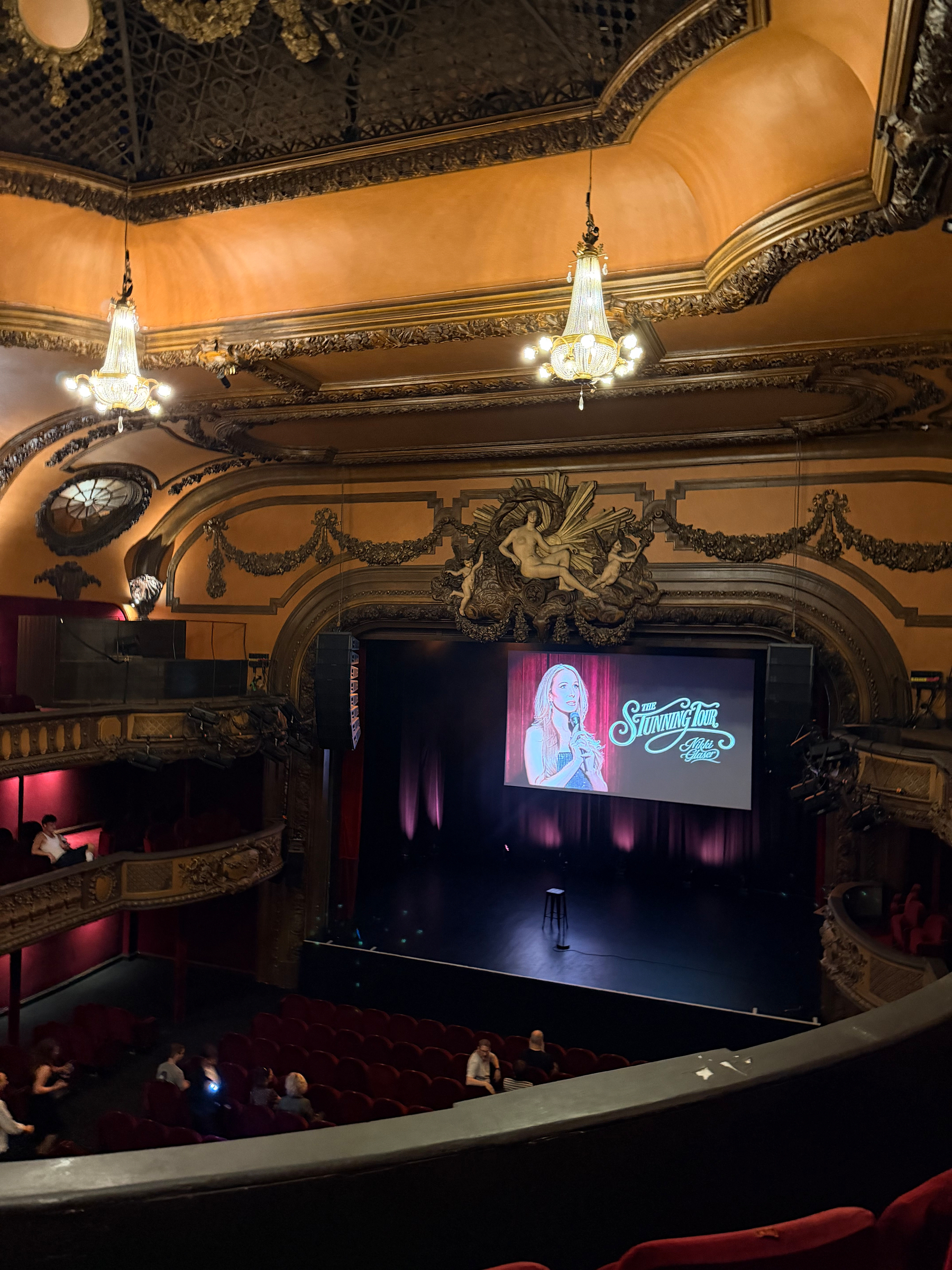 Auto-generated description: An ornate theater with a grand stage setup features a microphone stand and a backdrop promoting Strummy Time with Lizzie, as a small audience gathers.