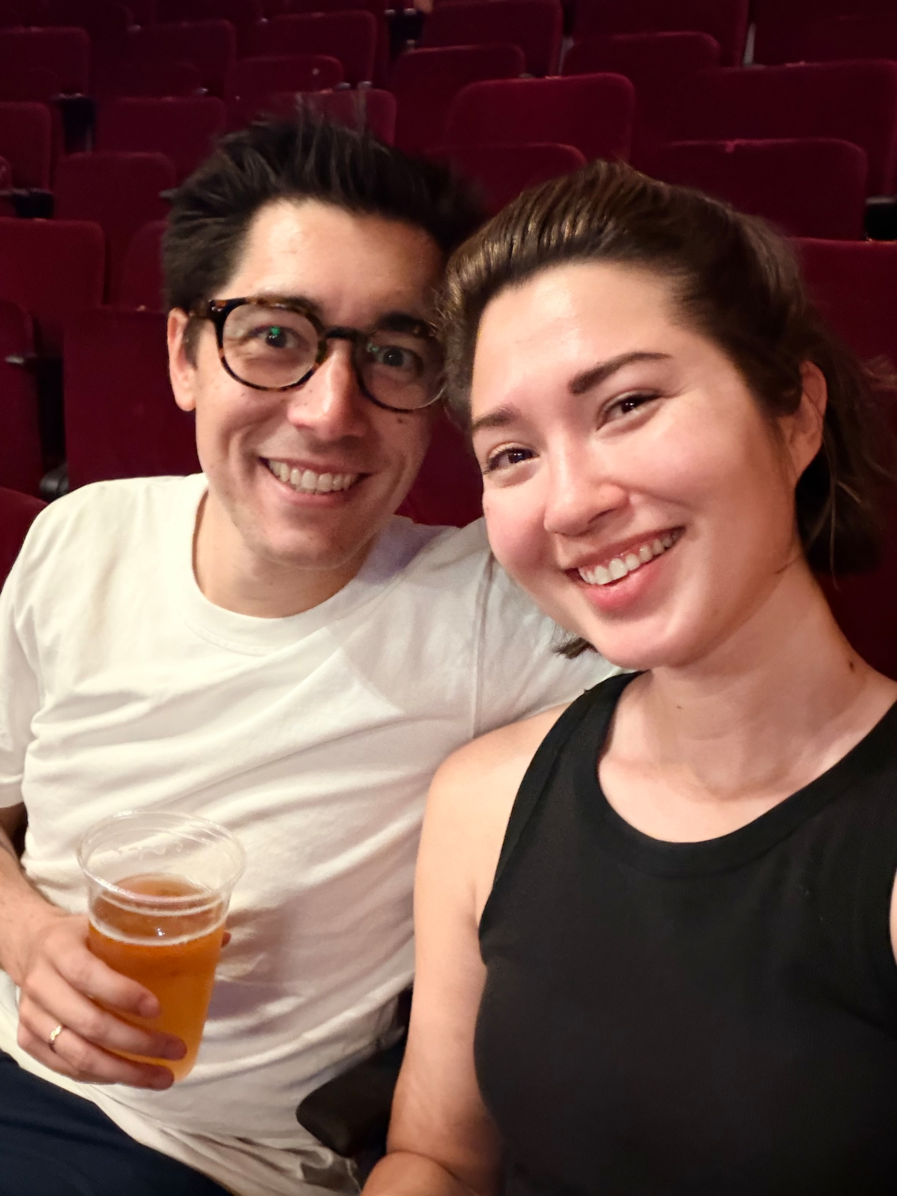 Auto-generated description: A smiling couple is seated in a theater or auditorium, with one holding a cup of beer.