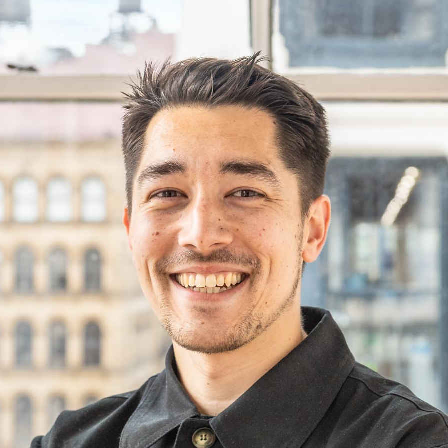 Auto-generated description: A smiling man with short dark hair and light facial hair is standing indoors with a blurred cityscape visible through the window behind him.